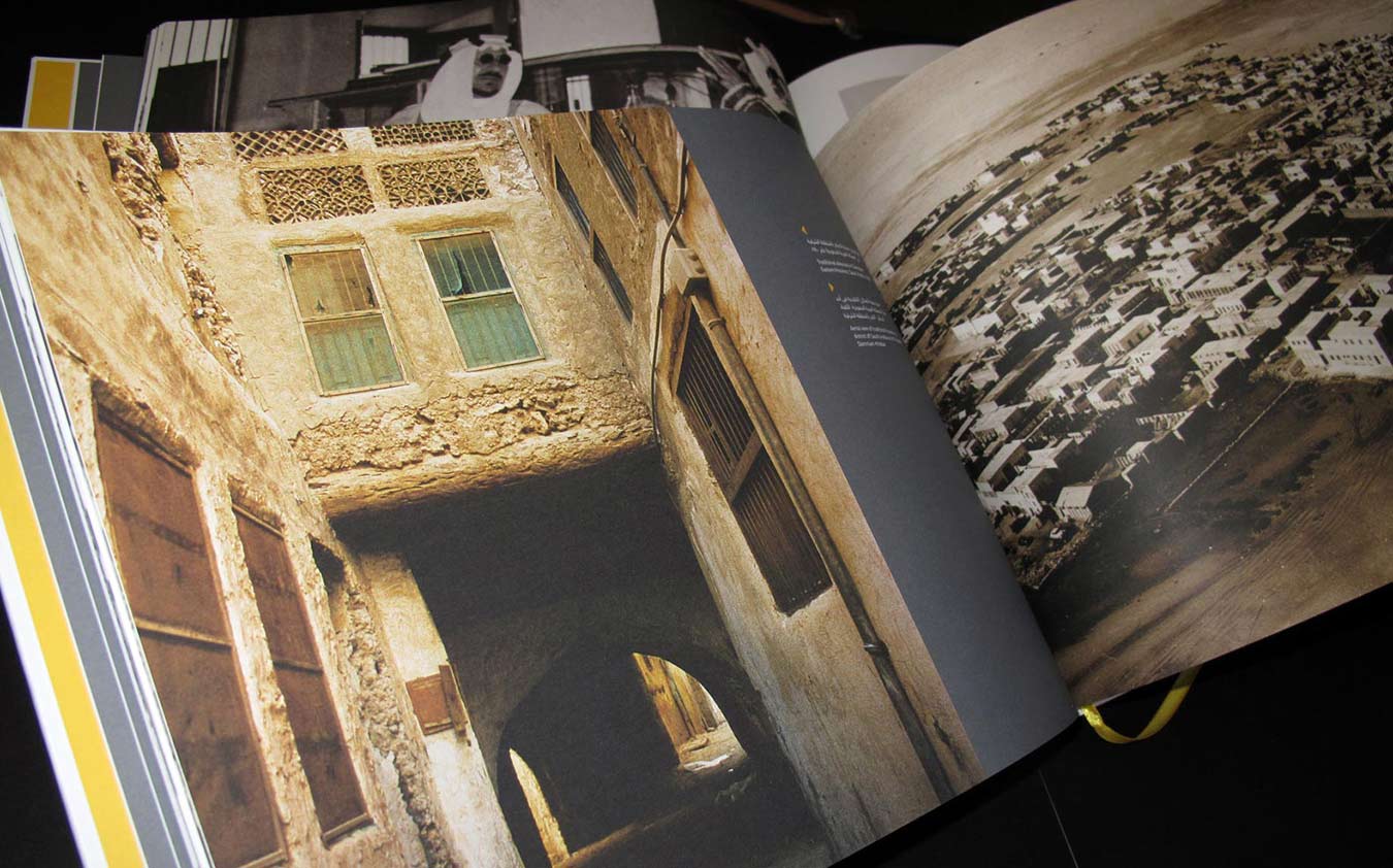 The open corporate landscape brochure spread displays an old, narrow alleyway with traditional stone arches and weathered facades on the left page. The right page features a historic, sepia-toned aerial photograph of a coastal town or settlement, creating a stark visual contrast that emphasizes heritage and historical development.