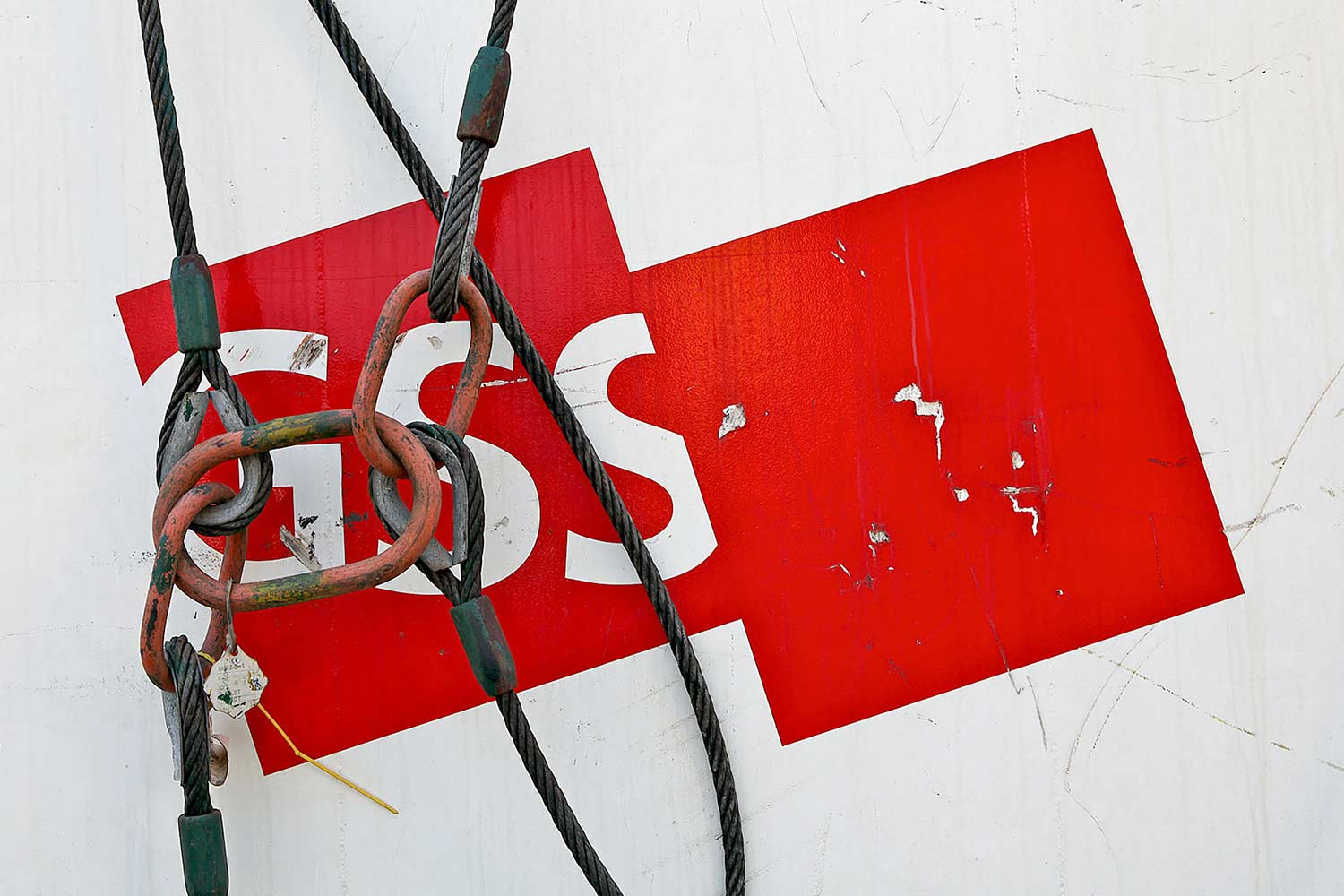 A bold red graphic shape with white lettering sits on a weathered white surface, heavily obscured and crossed by thick wire rigging cables and heavy, rusted metal chain links.