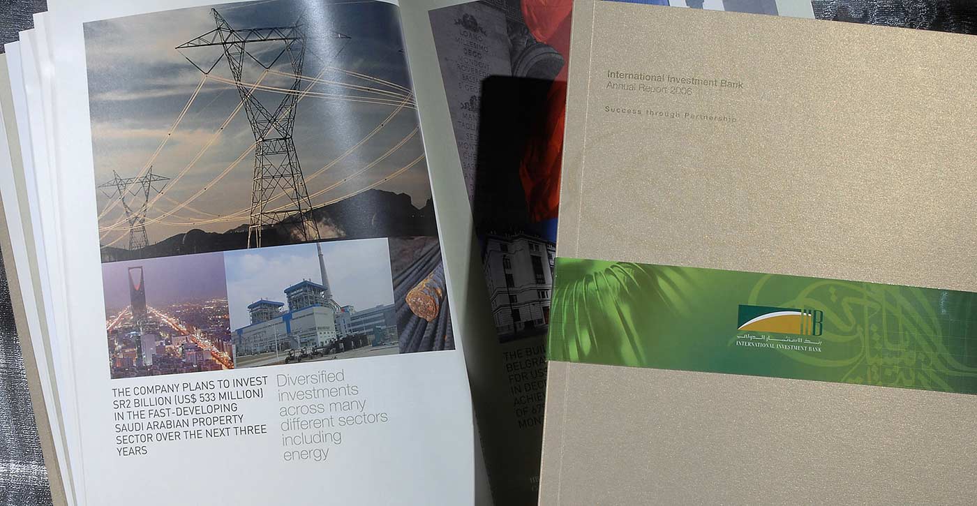 A close-up of open corporate publications for International Investment Bank, featuring an annual report with a gold cover and a green band, alongside an open page showcasing infrastructure photography and text regarding a 2 billion Saudi Riyal investment in the Saudi Arabian property sector.