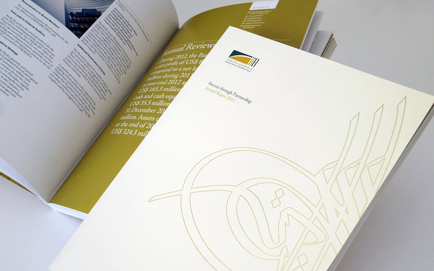 An open copy of the International Investment Bank Annual Report 2012, featuring a white cover with gold-colored linear calligraphy, the bank's logo, and the motto "Success through Partnership," with an open inner page showing financial review text in the background.