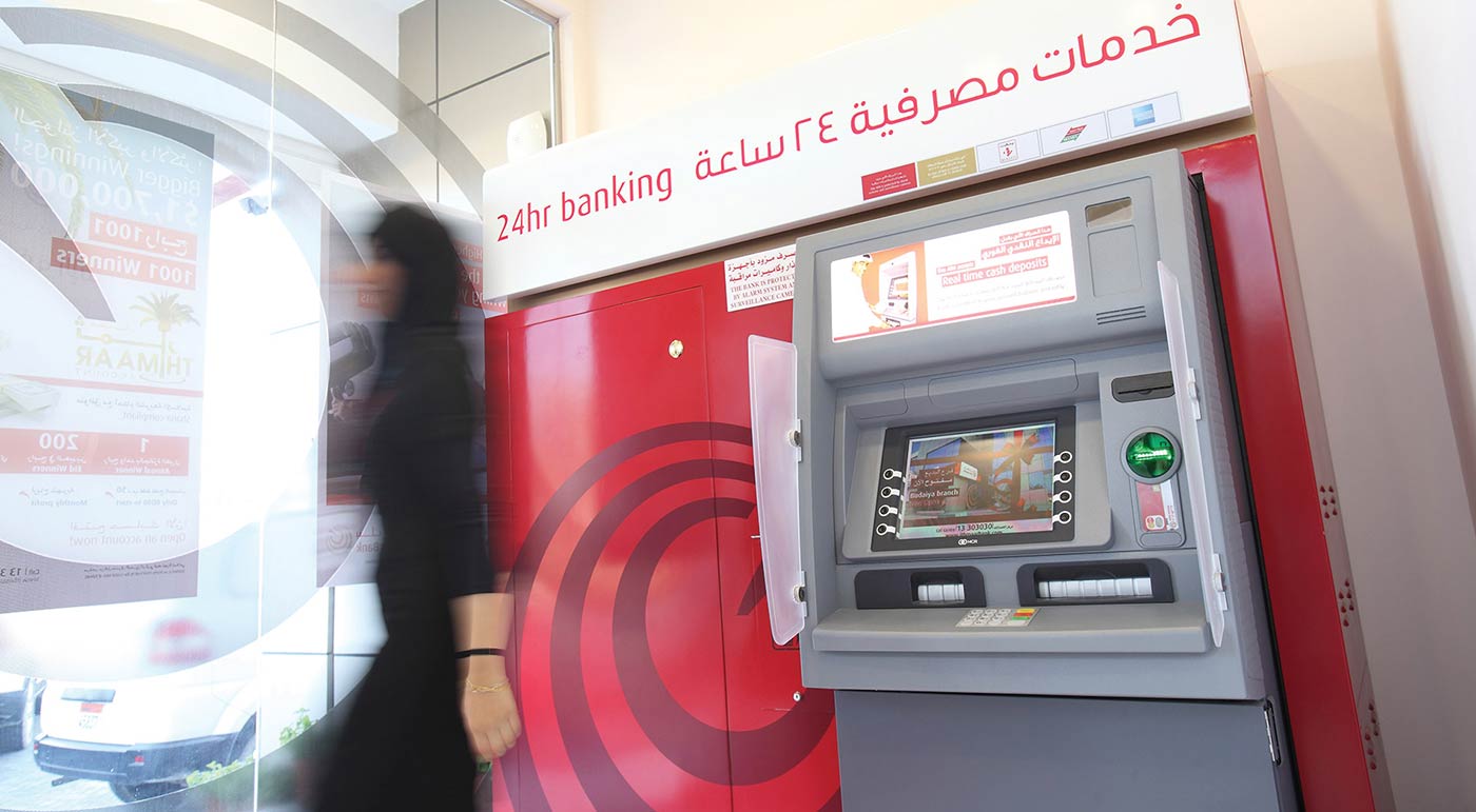 A person walking past an Ithmaar Bank 24-hour banking terminal, which is mounted on a red wall featuring the bank's circular logo design, with promotional signage visible in the background window.