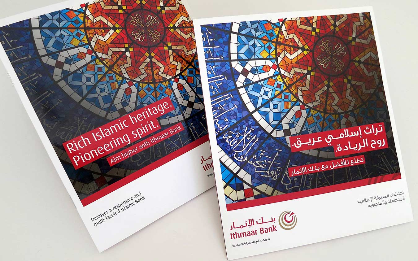 Two Ithmaar Bank promotional brochures lying side-by-side on a white surface. Both feature a vibrant, geometric stained-glass window design incorporating Arabic calligraphy. The left brochure displays English text reading "Rich Islamic heritage. Pioneering spirit." and "Aim higher with Ithmaar Bank," while the right brochure displays the equivalent promotional messaging in Arabic, along with the bank's official logo.