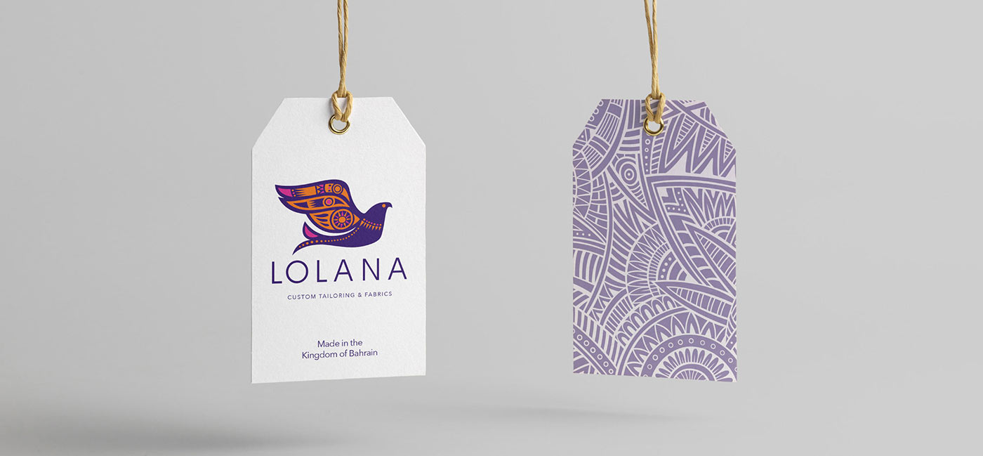 Two hanging clothing hangtags for LOLANA Custom Tailoring & Fabrics, displaying a white front tag with the colorful bird logo and "Made in the Kingdom of Bahrain" text next to a back tag featuring an intricate purple tribal pattern.