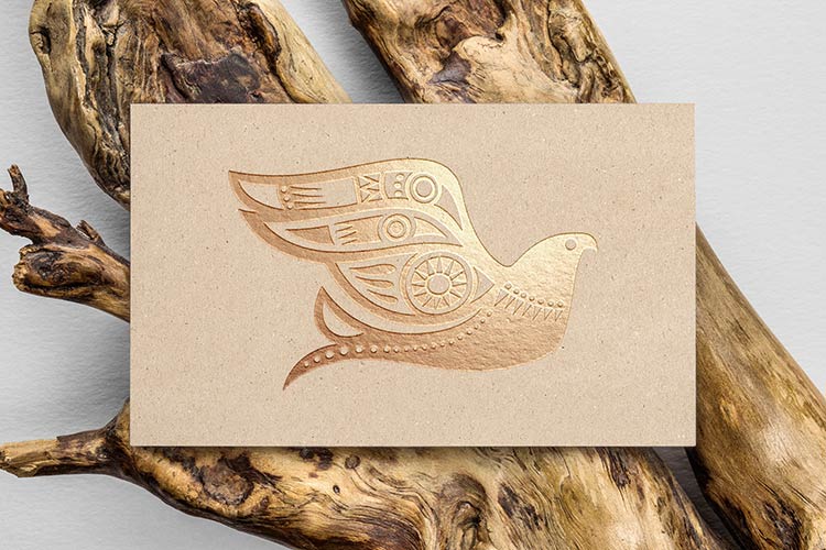 A kraft paper business card featuring a stylized bird logo hot-stamped in metallic gold ink, resting on a textured piece of natural driftwood.