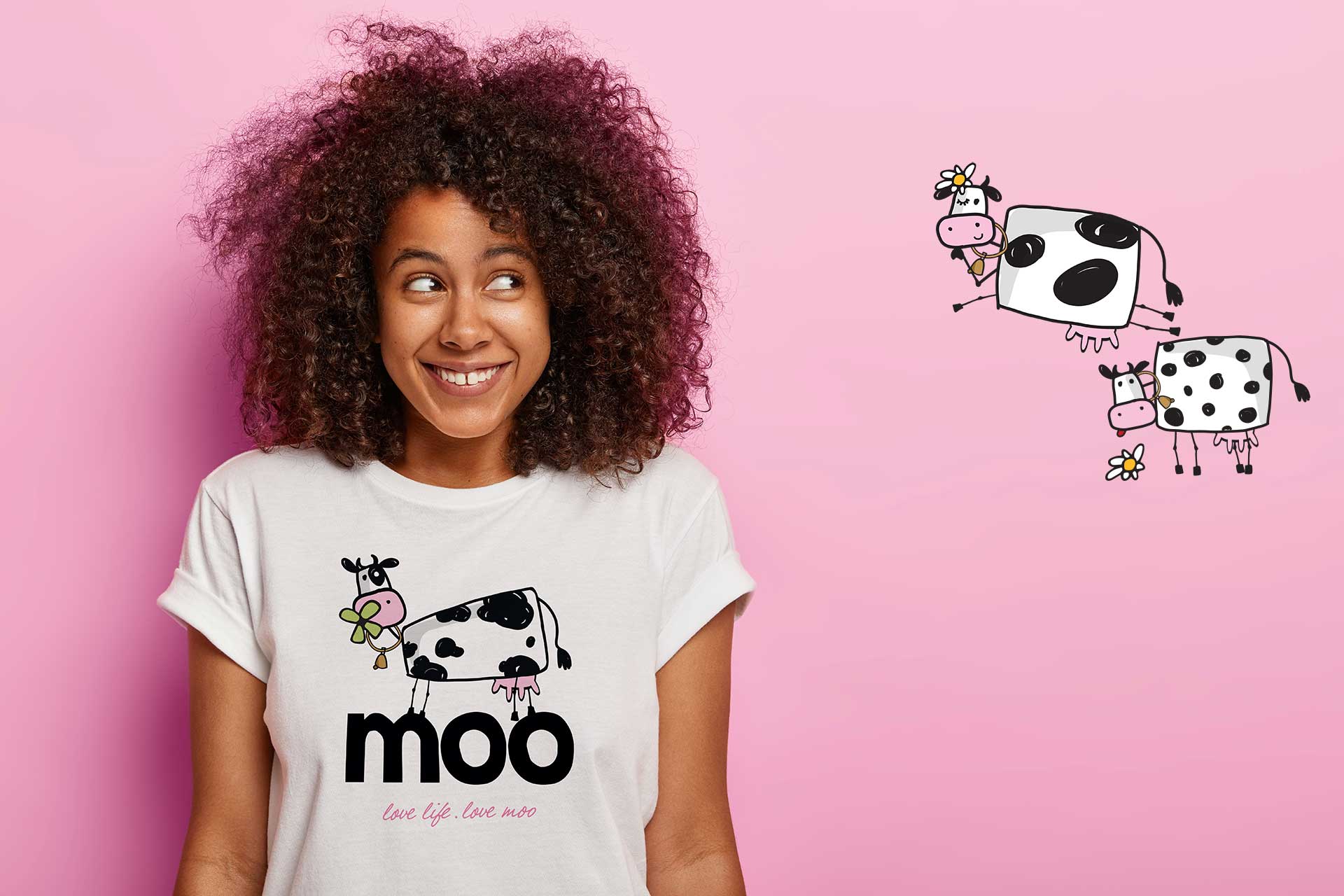 A smiling young woman with curly hair wears a white graphic t-shirt featuring a whimsical cartoon cow and the word "moo" against a bright pink background.