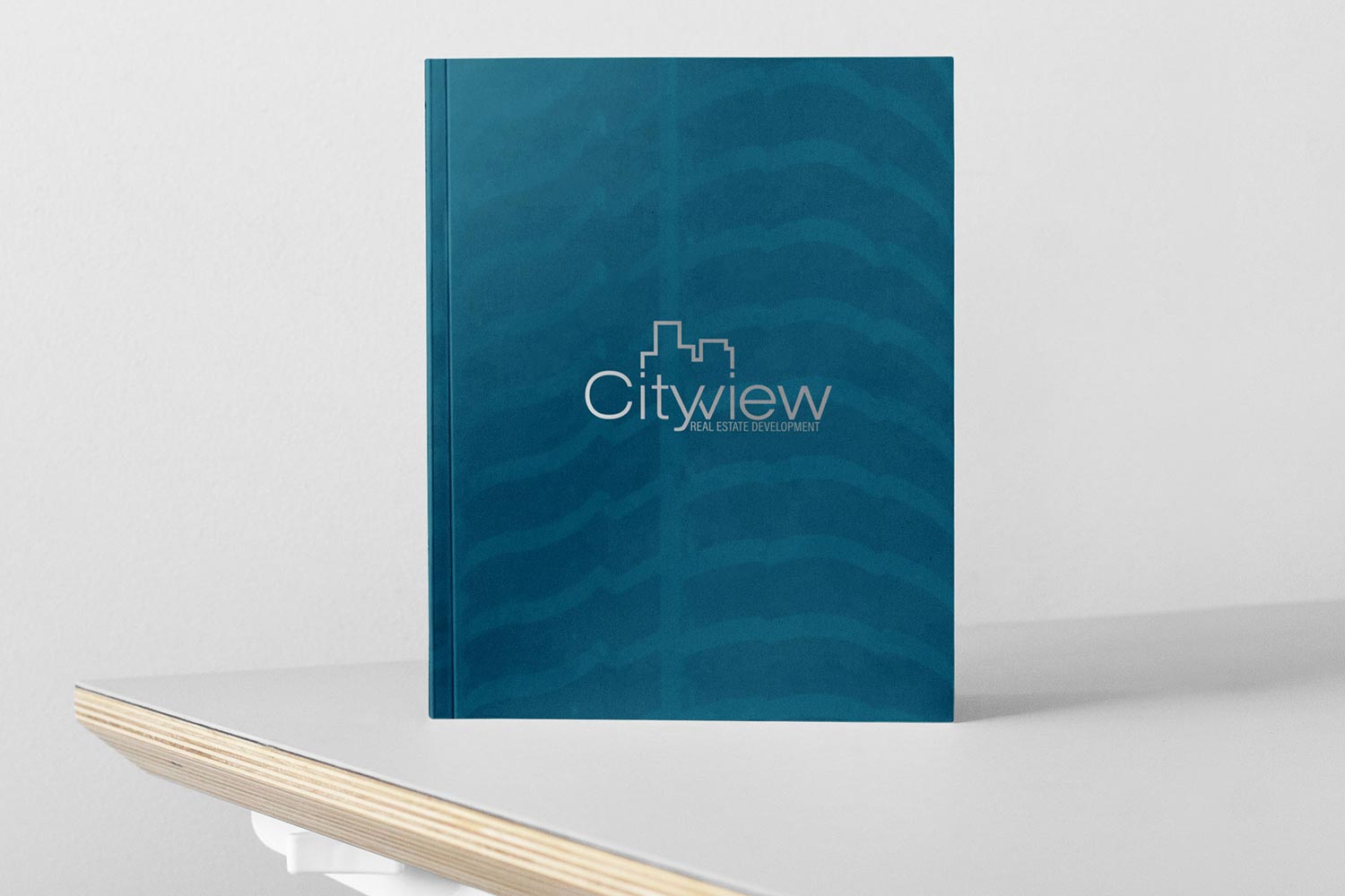 A closed teal-blue Cityview Real Estate Development brochure featuring a silver metallic logo on its subtly textured cover, standing upright on the edge of a white table.