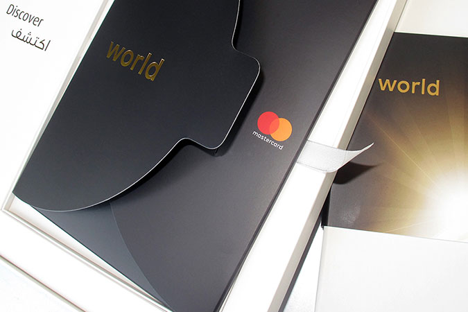 A premium presentation package for a "World" Mastercard, featuring a matte black folder with gold "world" lettering, a white ribbon, and an adjacent companion brochure with a sunburst design, all set against a clean, white background with the word "Discover" printed in both English and Arabic.