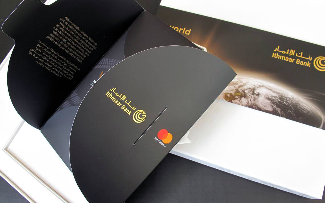 An open black card presentation folder from Ithmaar Bank, featuring gold branding, a Mastercard logo, and English and Arabic text describing the benefits of a "World" contactless credit card, alongside a partial view of a planet Earth graphic on the inner panel.