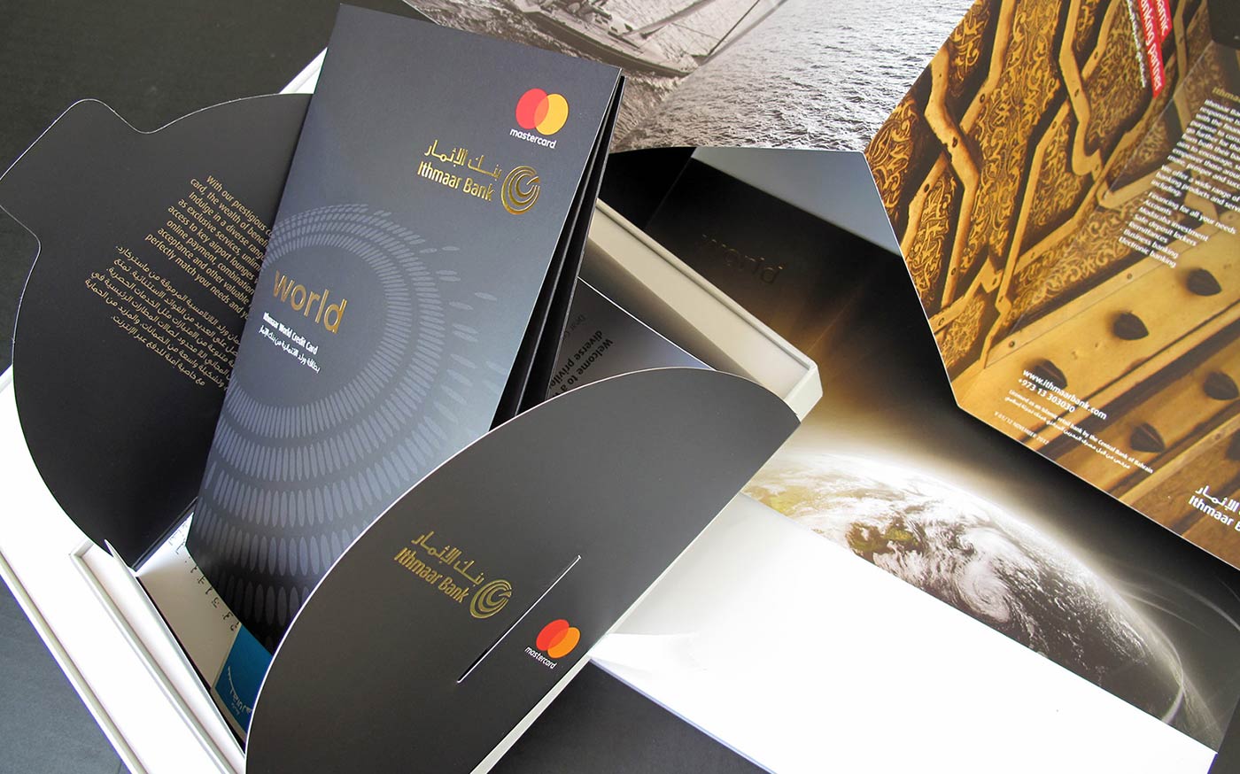 An open black card presentation folder from Ithmaar Bank, featuring gold branding, a Mastercard logo, and English and Arabic text describing the benefits of a "World" contactless credit card, alongside a partial view of a planet Earth graphic on the inner panel.