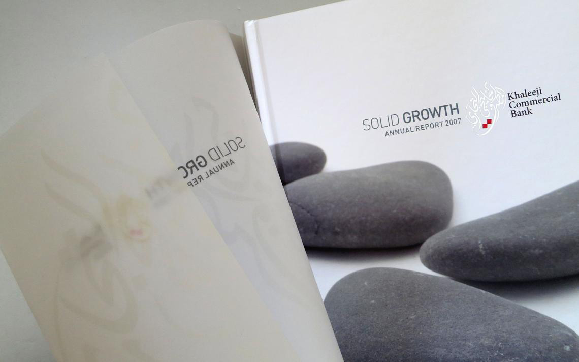 the white cover of the Khaleej Commercial Bank 2007 Annual Report, titled "SOLID GROWTH." The design features smooth, dark gray river stones in the foreground. On the left side of the frame, a semi-transparent vellum or tracing paper flyleaf page is partially turned, showing a mirrored, faint reflection of the title text from the underlying page.