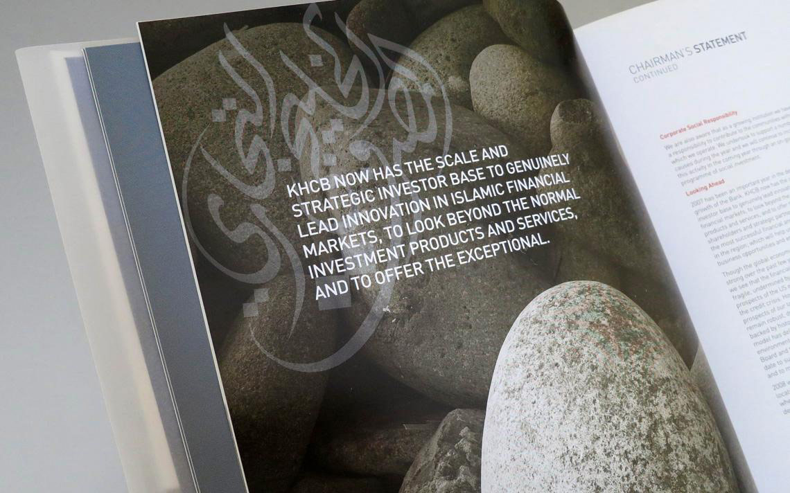 an open spread of a corporate report titled "CHAIRMAN'S STATEMENT CONTINUED." The left page features a full-bleed photograph of smooth, stacked river stones overlaid with large white text about KHCB's strategic investor base and Islamic financial markets, layered with faint Arabic calligraphy. The right page displays editorial text sections under headings like "Corporate Social Responsibility" and "Looking Ahead," with a large, textured light-colored pebble partially curving into the frame from the bottom.