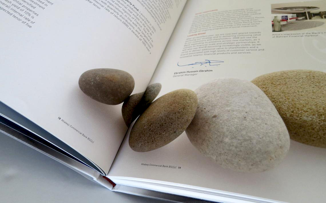 shows an open annual report or corporate brochure for Khaleej Commercial Bank. Several smooth, rounded river stones or pebbles are arranged decoratively across the center fold of the pages. The right page features a message signed by Ebrahim Hussain Ebrahim, the General Manager, alongside corporate text columns and a small architectural rendering in the top right corner labeled "Artist's impression of the Bank's room at Bahrain Financial Harbour."